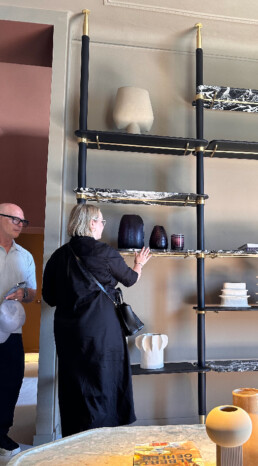 Two people look and touch a tall open-style shelving unit against a wall with decor sitting on it's shelves.