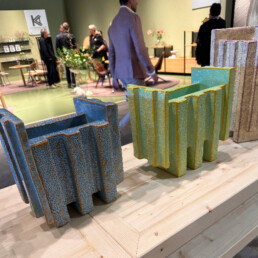 Three geometric vases sit on a shelf. Behind the shelf are people walking around at the Salone del Mobile fair.