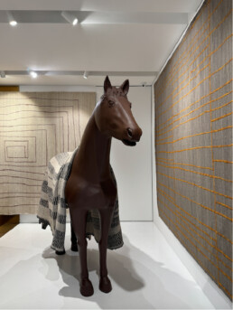 A statue of a horse sits between two carpets hanging on two walls. The horse statue has a rug on it's back as well. These three rugs are all part of the newest Golran collection release by Pieor Lissoni called Plaid. The rugs are mostly simple, with small irregular lines on them in geometri]c shapes.