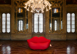 The Gufram Bocca lips sofa sits at the center of a room, seemingly housed within a centuries-old building. The room is embellished with a grand hanging chandelier, mirrors featuring golden embossed frames, and ornate walls, windows, and floors.