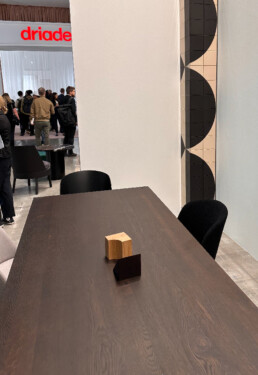 A dining table sits centre reaching the bottom of the photo. The table has two decorative accessories sitting on stop. In the background are people walling and standing around at Salone del Mobile furniture fair.
