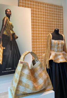 The image showcases the latest release from Italian rug makers, Kasthall: the Vichy rug. With its classic checkered design, this particular rug comes in a washed-out yellow and light beige color scheme. The image captures three main focal points: the primary rug hanging from the ceiling in the background, a mannequin adorned with a shirt and purse made from the same fabric, and a poster featuring a model wearing the same shirt and holding the same bag, accompanied by the text 