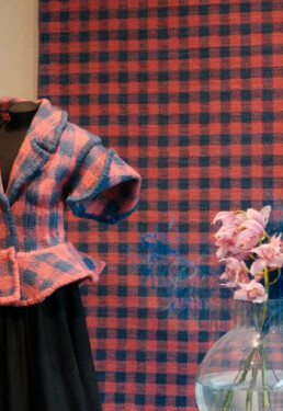 A single piece of designed fabric drapes over two spaces: first, adorning a mannequin dressed in a beautiful jacket, and second, hanging behind the mannequin as a decorative rug. The design features a classic checkered pattern in shades of pinkish red and washed-out navy blue. There is also a vase with flowers in the bottom right corner of the image.
