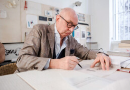 A man (Piero Lissoni) sits at a table with a pen and paper drawing, what we can presume, are furniture designs. He wears glasses and a suit jacket with a dress shirt looking professional yet artisitic.
