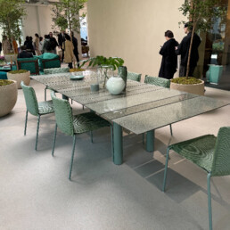A room filled with plants, people walking around, and a large beautiful aqua-coloured outdoor dining table mixed of small, medium and large horizontally placed glass slats. The table is surrounded by 6 chairs in the same colour, though in a more plush fabric material.