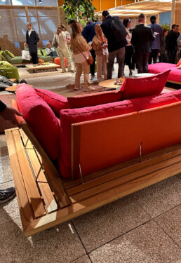 A section of the back of a large seating system in the middle of a living space (the Roda exhibition at the Rho Fieramilano fairgrounds) is highlighted, showcasing its clean teak frame and bright pink fabric padding.