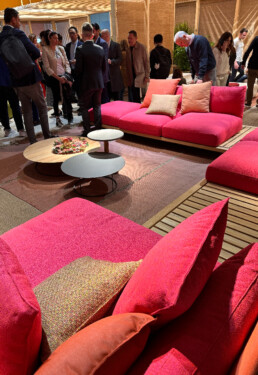 A large seating system in the middle of a living space (the Roda exhibition at the Rho Fieramilano fairgrounds) is highlighted, showcasing its clean teak frame and bright pink fabric padding.