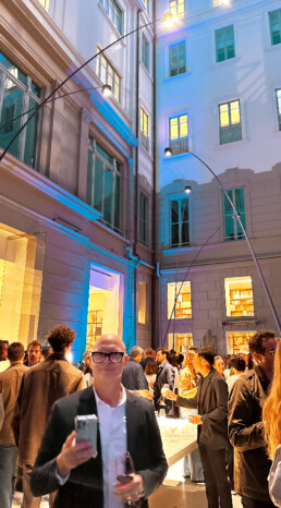 Ross Bonetti, CEO of Livingspace, attends the Davide Groppi party in Milan. The photo is set in a large courtyard adorned with massive Davide Groppi lights, spanning from the floor to four meters above the crowd of people chatting and enjoying drinks.