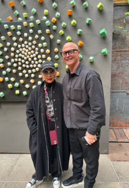 In the company of the renowned Italian artist, Rossana Orlandi, stands Ross Bonetti, a distinguished businessman hailing from Vancouver. They are pictured together in a room adorned with an intriguing art installation made by Rossana as their backdrop.