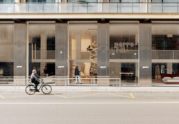 A bank bikes past a store front, while a woman steps inside. The storefront has five huge paning glass windows - one of which is a door. Between the large pans of glass is a metal facade with the brand name 