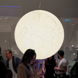A large light dominates the foreground of the image, with people walking around, relaxing, and socializing nearby. The circular light appears to be wrapped in thin, carefully crafted paper, giving it a light and slightly translucent design.