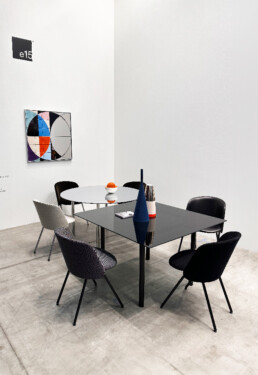 The image highlights a dining set from the furniture brand e15. There are two small dining tables, one circular and one square, 6 chairs total circle around the two tables. There is a unique piece of geometric art on the walls and the e15 logo.